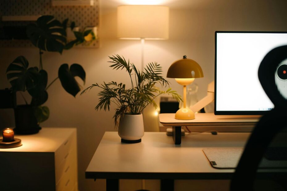 Mesa de trabalho iluminada com luminária retrô, planta decorativa e monitor, em ambiente acolhedor e organizado para home office.