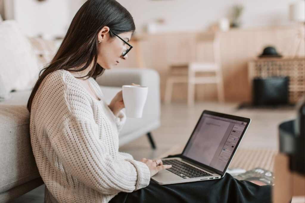 Mulher trabalhando em laptop no chão ao lado do sofá, com xícara de café, em um ambiente de trabalho flexível em casa.