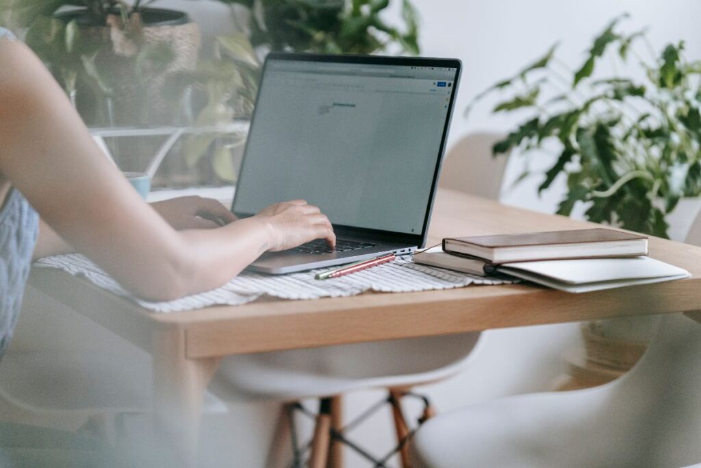 Pessoa trabalhando em laptop em uma mesa de madeira clara com plantas ao fundo, destacando um ambiente de trabalho doméstico organizado e iluminado.
