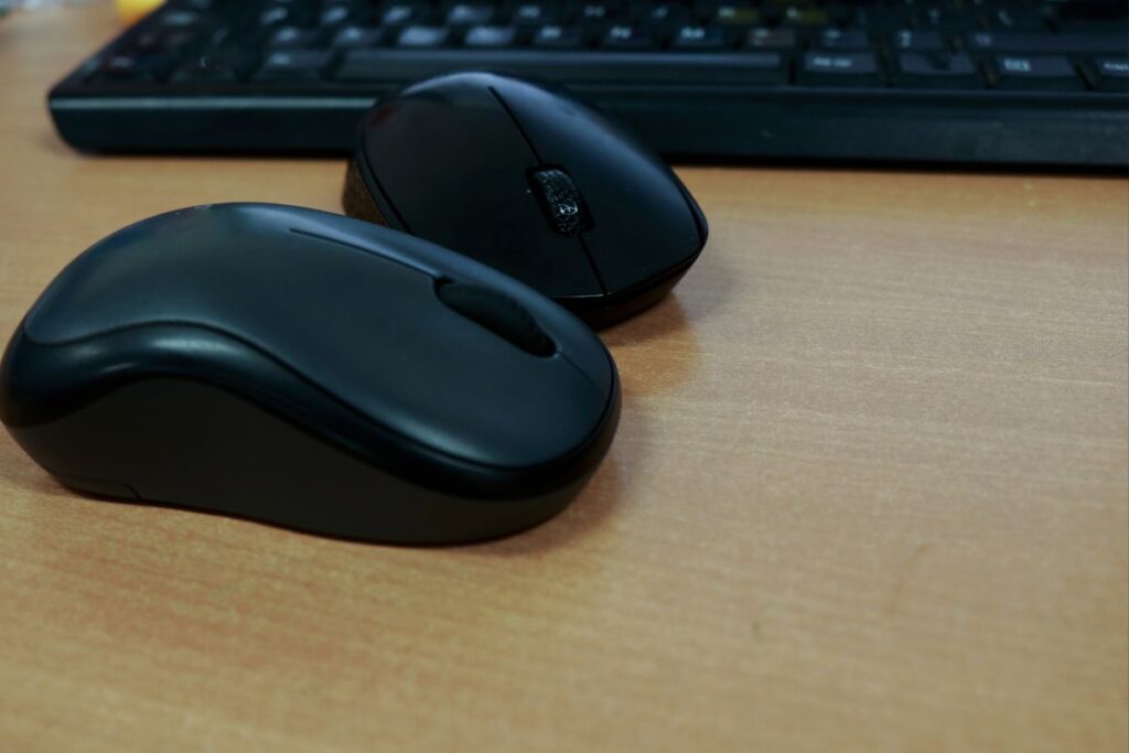 Dois mouses sem fio pretos sobre uma mesa de madeira clara, com um teclado desfocado ao fundo, ideal para flexibilidade no trabalho.