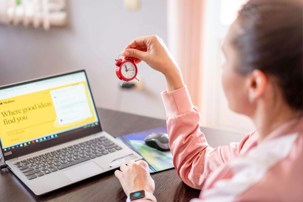Mulher segurando mini despertador vermelho em frente ao notebook aplicando o método pomodoro de intervalos de foco