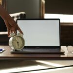 Mão ajustando despertador branco sobre mesa com notebook e óculos representando a técnica pomodoro de gestão do tempo