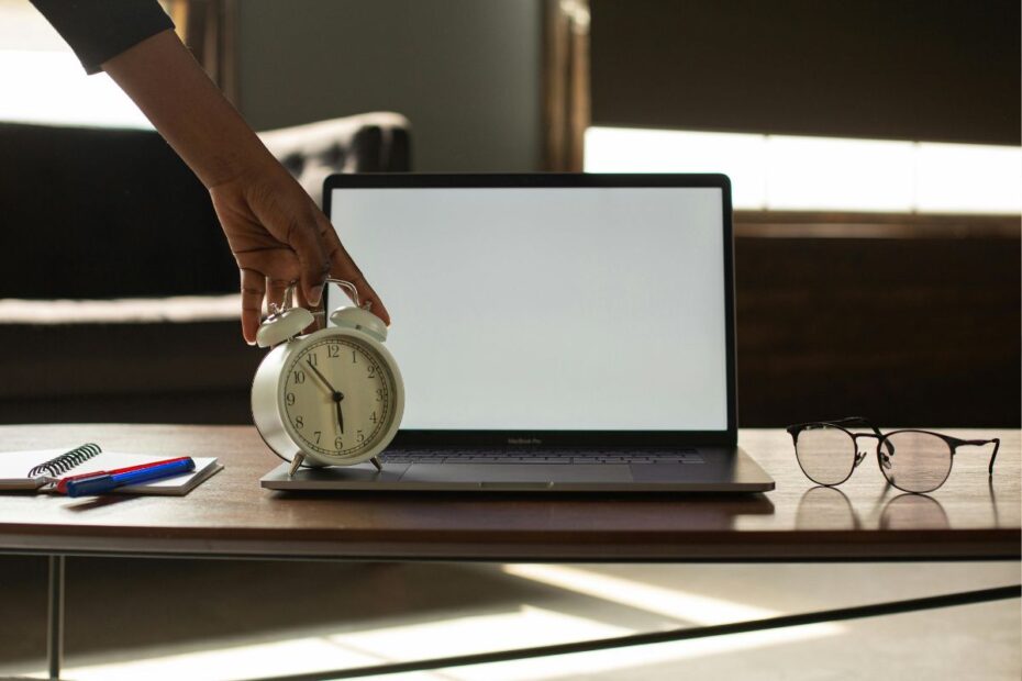 Mão ajustando despertador branco sobre mesa com notebook e óculos representando a técnica pomodoro de gestão do tempo